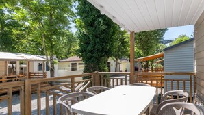 A covered outdoor area with a table and chairs, surrounded by wooden railings and trees. - Mobile Home 'Confort A - 1-8 Pers' with Shared Pool, Private Terrace and Shared Garden (Lavilledieu)