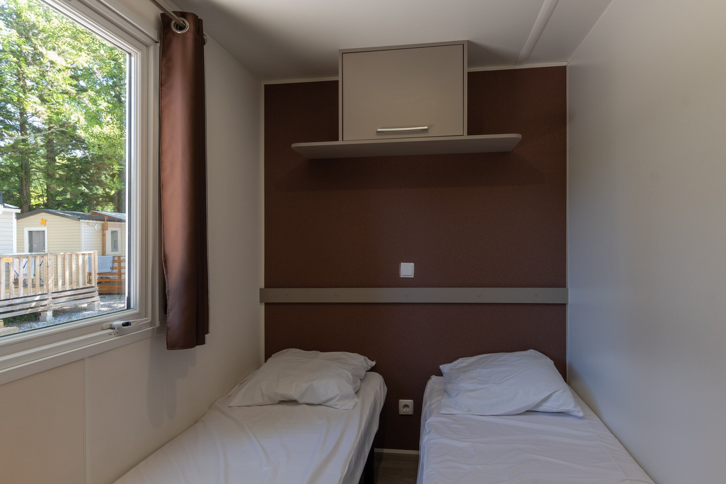 Une petite chambre d’hôtel composée d’une seule pièce, avec deux lits, une fenêtre avec des rideaux, une étagère intégrée au-dessus des lits et une vue sur un quartier résidentiel.