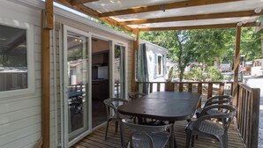 A covered outdoor deck with a table and chairs, a sliding glass door leading to a kitchen, and a view of a residential area. - Mobile Home 'Mobil-home Confort B - 1-8 Pers' with Shared Pool and Shared Garden (Lavilledieu)