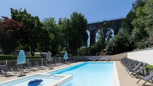 A swimming pool with lounge chairs and umbrellas, a bridge in the background, and trees around. - Mobile Home 'Mobil-home Confort B - 1-8 Pers' with Shared Pool and Shared Garden (Lavilledieu)