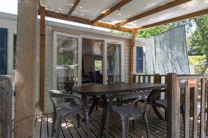 Ett takförsett utomhusområde med ett bord och stolar, träbjälkar och utsikt över ett hus med glasdörrar.