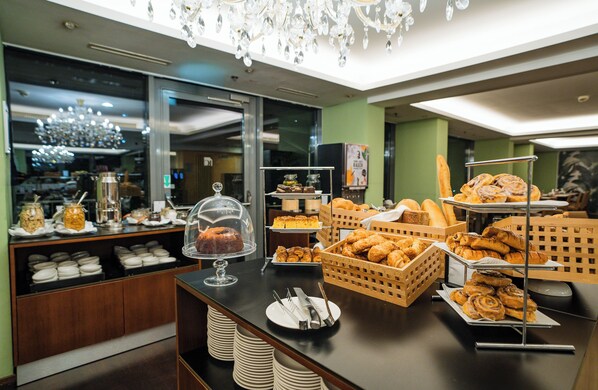 Un bufé con diversas pastelerías y postres, entre ellos, cruasanes y pasteles, expuestos en una barra con utensilios para servir.