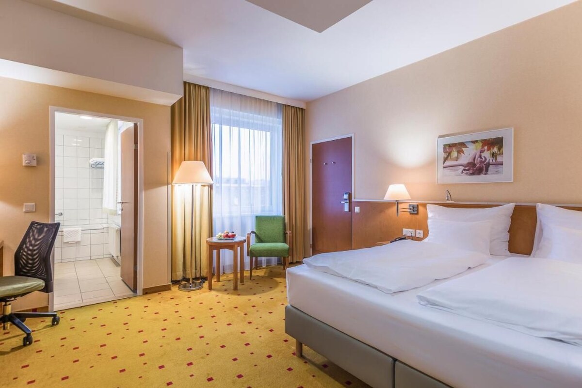 Een hotelkamer met een bed, een stoel, een klein tafeltje met een lamp, een deur en een raam met gordijnen.