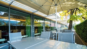 Een buitenterras met tafels en stoelen onder een overkapping, omringd door glazen wanden en een haag.