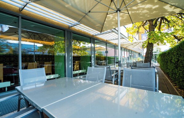Un espacio de comedor al aire libre con mesas y sillas bajo una cubierta, rodeado de paredes de vidrio y un seto.