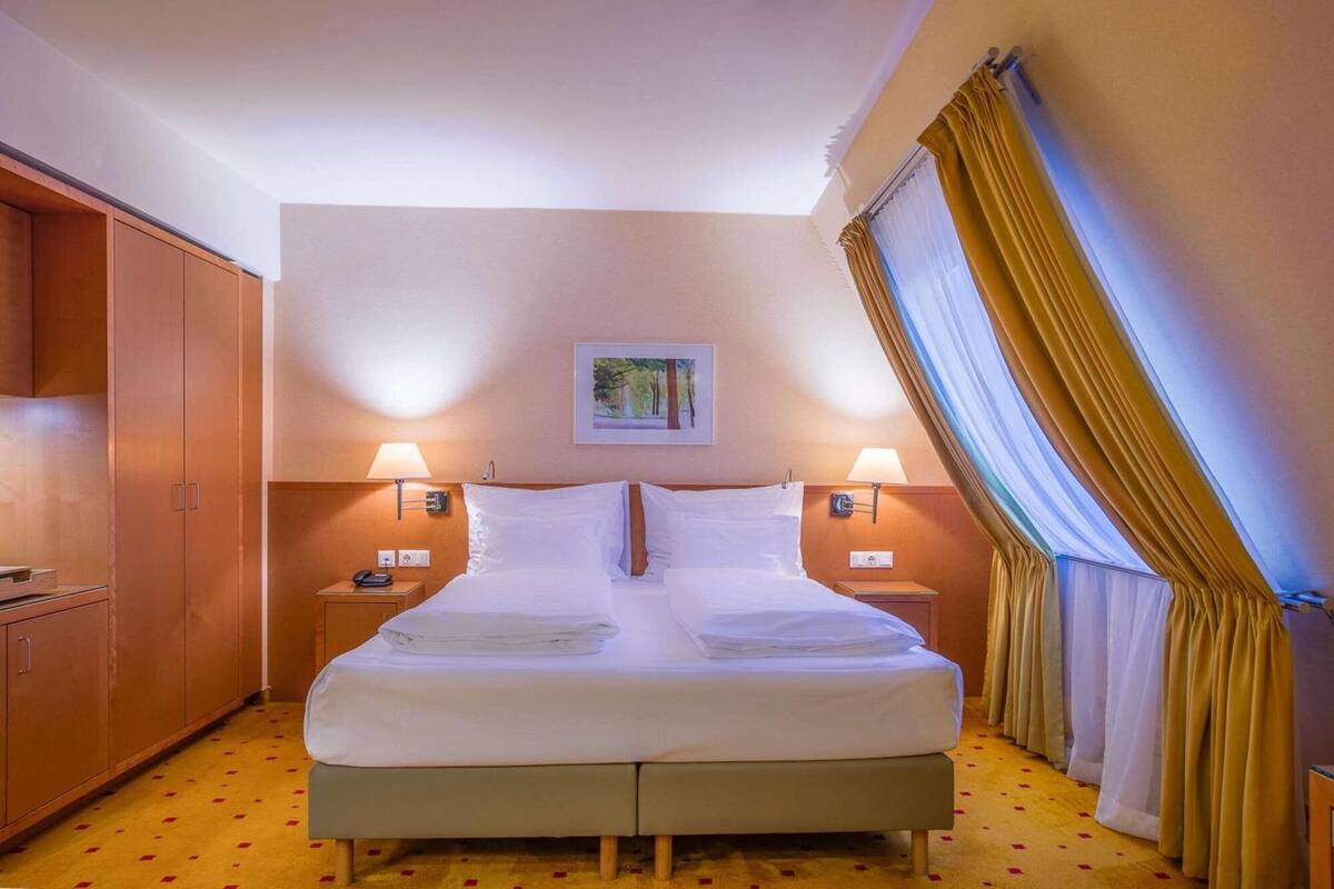 A hotel room with a double bed, two bedside tables with lamps, a wooden wardrobe, and a painting on the wall.