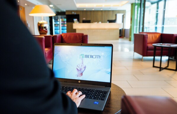 Una persona usando una computadora portátil en un moderno vestíbulo de hotel con asientos rojos y una recepción al fondo.