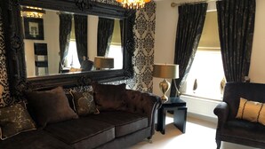 A dark-colored sofa with multiple cushions, a small black side table with a lamp, a large ornate mirror, and a dark armchair. - Sheffield Hillside Retreat (Sheffield)