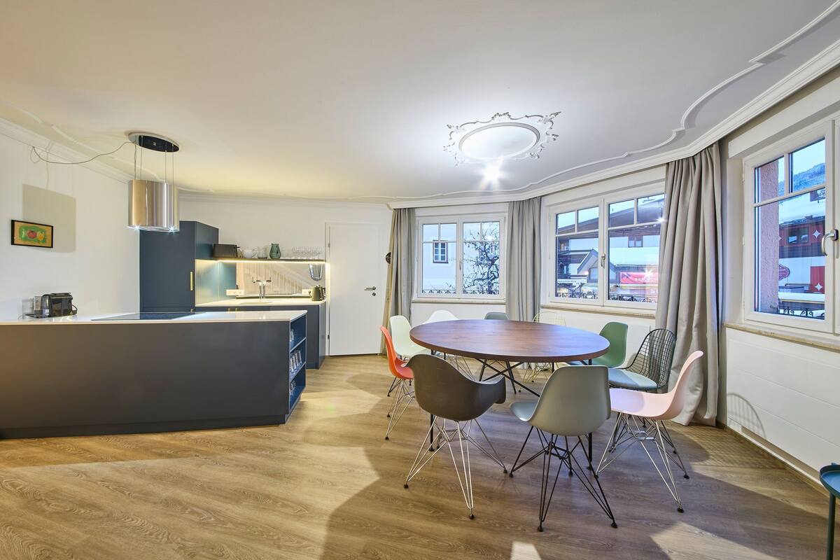 A modern kitchen with a central island, a dining area with a round table and colorful chairs, and large windows offering a view of the outside.