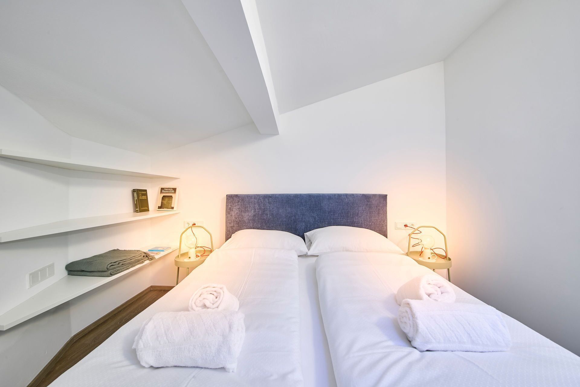 Una habitación de hotel con una cama, dos mesitas de noche, una estantería con libros y un cabecero.