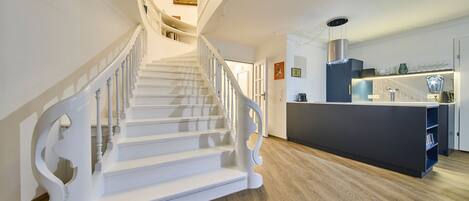 A staircase with white balusters leading to a landing with a painting. A kitchen area with a dark blue island and modern appliances is visible.