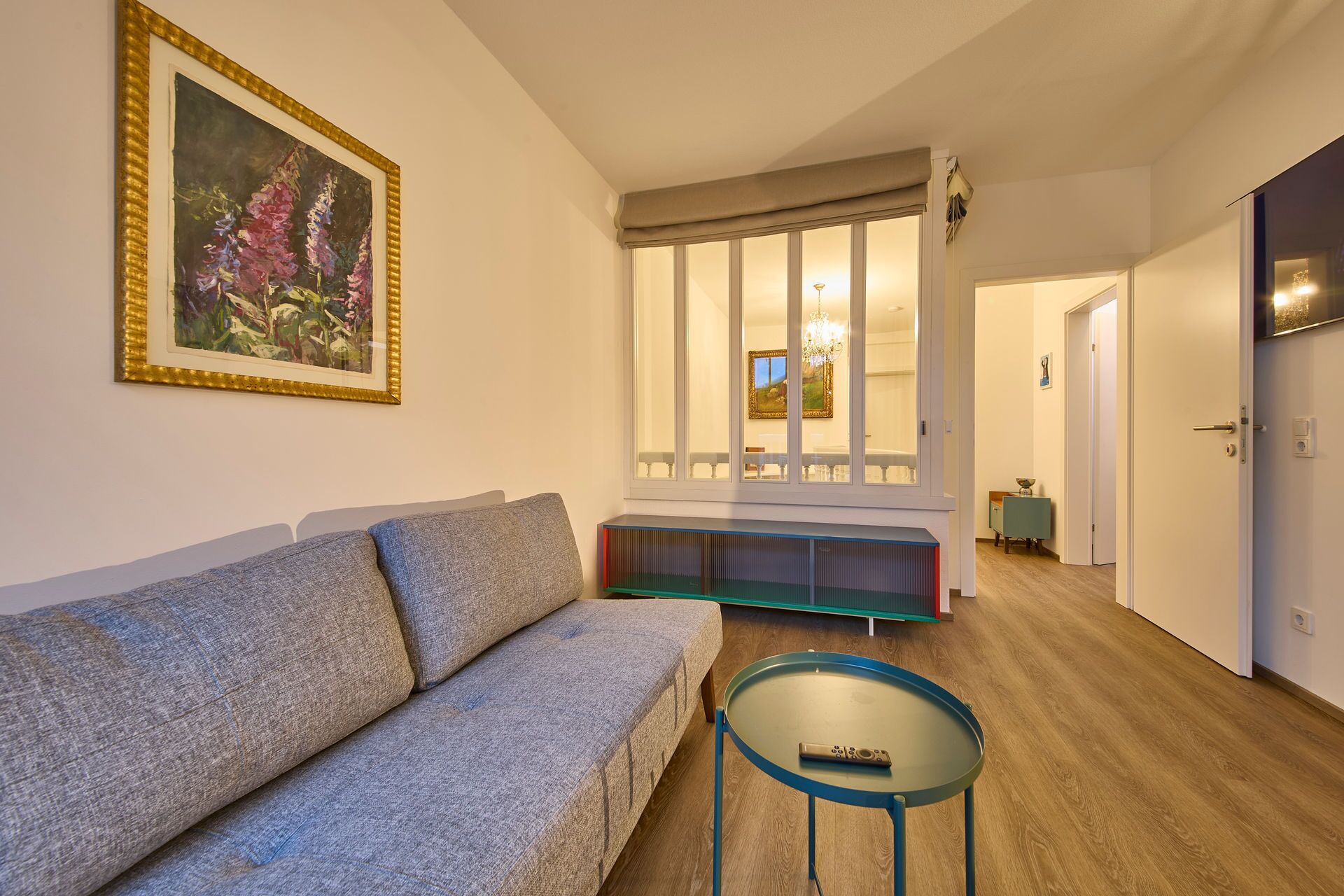 A living room with a sofa, a glass coffee table, a painting, a bench, and a view through a window.