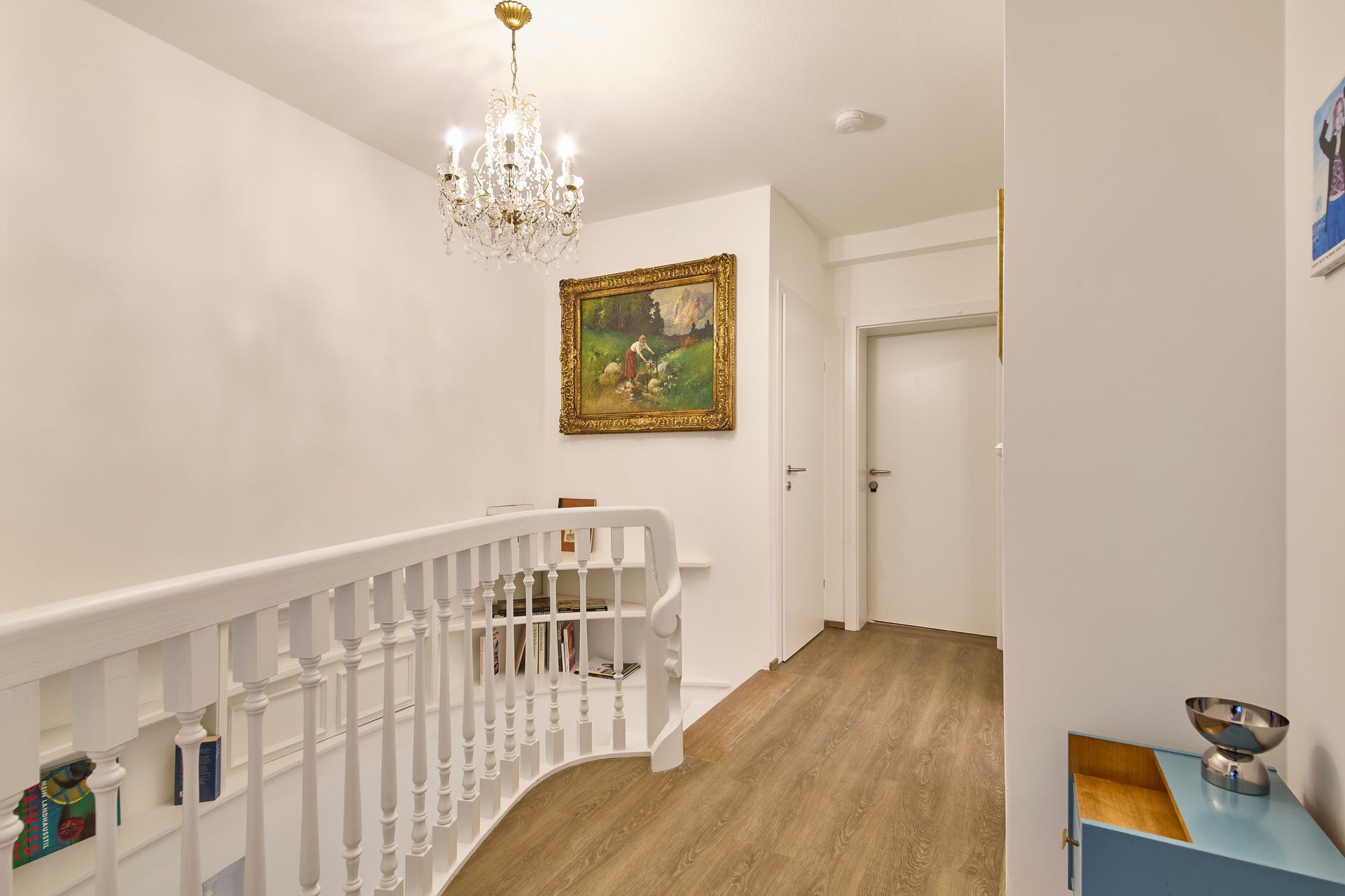 Un couloir avec une rambarde blanche, un lustre, un tableau accroché au mur et une porte au fond.
