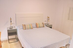 A bedroom with a bed, two bedside tables, a nightstand, a lamp, and a wall socket.
