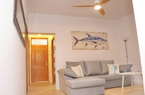 Un salon avec un canapé gris, une porte en bois, un ventilateur de plafond et un tableau représentant des poissons accroché au mur.