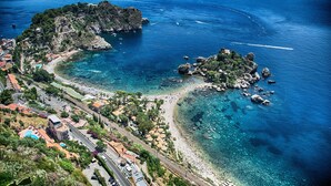 An aerial view of a coastal town with clear blue waters, rocky outcrops, and a mix of residential and commercial buildings. - SMERALDO (Giardini Naxos)