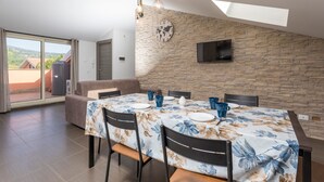 A dining area with a table set for four, a stone wall, a clock, and a TV mounted on the wall. - Apartment 'Sophia' with Mountain View, Shared Terrace and Air Conditioning (Fiumefreddo Sicilia)