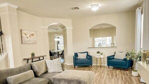 Un salon avec un canapé beige, un fauteuil bleu et un pouf assorti. On y trouve une petite table ronde avec une plante, et un coin repas avec une table et des chaises est visible à travers une arche.