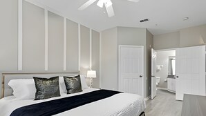 Une chambre avec un grand lit, une table de chevet, un ventilateur de plafond et une porte donnant sur une salle de bain.