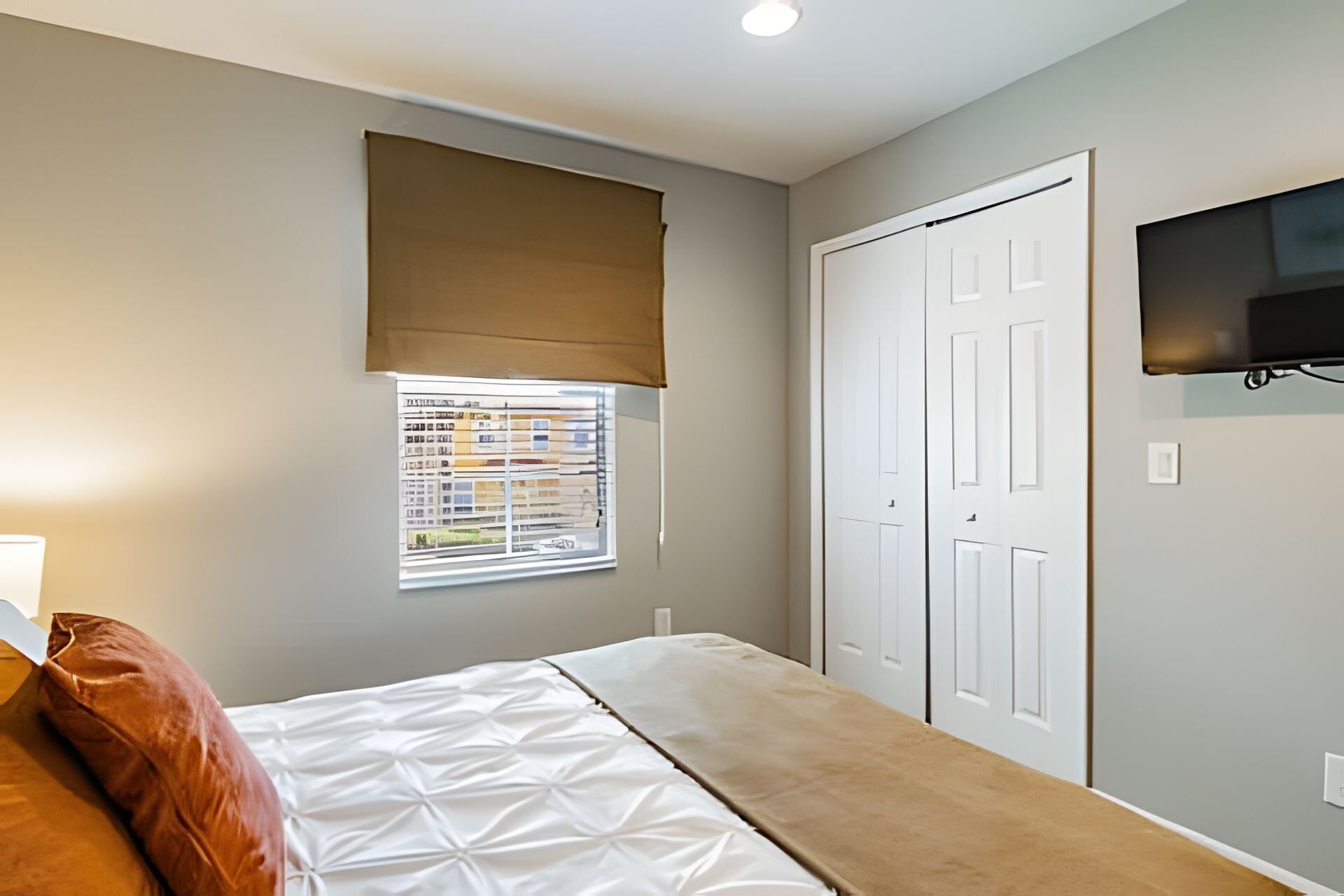 Une chambre à coucher avec un lit, une fenêtre avec des stores, une armoire et un téléviseur fixé au mur.