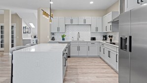 A modern kitchen with white cabinets, a large island, stainless steel appliances, and a pendant light fixture. - Windsor Hills 6BR Home, Private Pool + Game Room (Kissimmee)