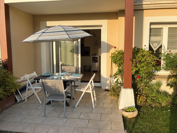 Een terras buiten met een tafel gedekt voor twee personen, een gestreepte parasol en een tuin met verschillende planten en bloemen.