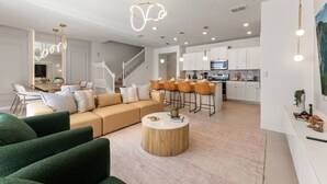 Una sala moderna con un sofá, una mesa de centro y sillas verdes. Cuenta con una zona de cocina con armarios blancos y un comedor con taburetes de bar.