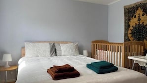 Un lit bien fait, avec une couette blanche, deux oreillers et une couverture marron pliée. On y voit un berceau avec un cadre en bois et un matelas blanc. Une lampe est posée sur une table de chevet, et une tapisserie est accrochée au mur.