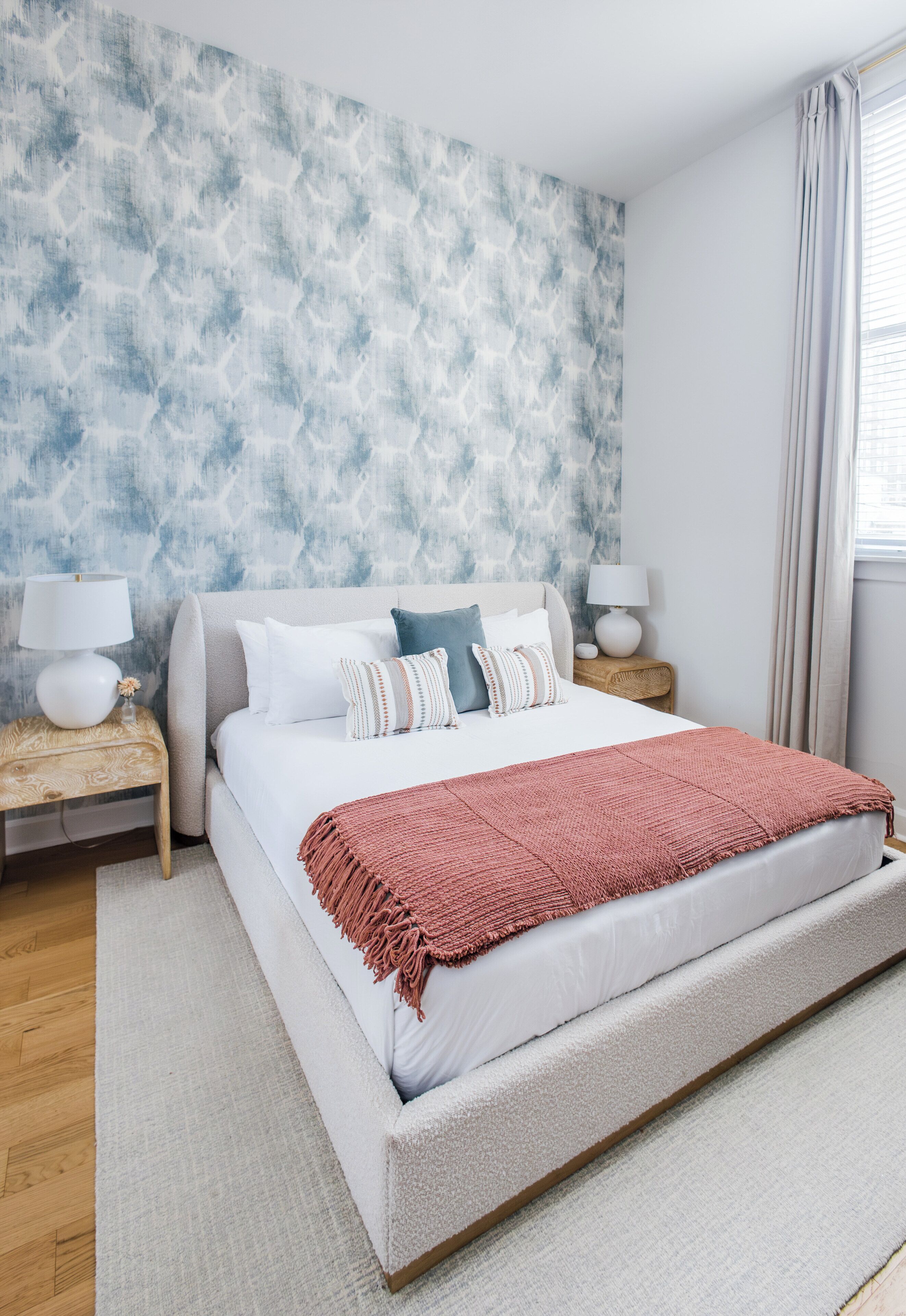 A bedroom with a large bed, two bedside tables with lamps, a patterned wall, and a window with curtains.