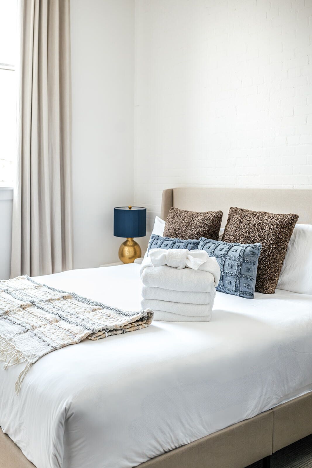 Una cama bien hecha con una colcha blanca, una toalla blanca doblada y una manta texturizada. Hay dos almohadas con un estampado marrón y azul, y una lámpara azul sobre una mesita de noche.