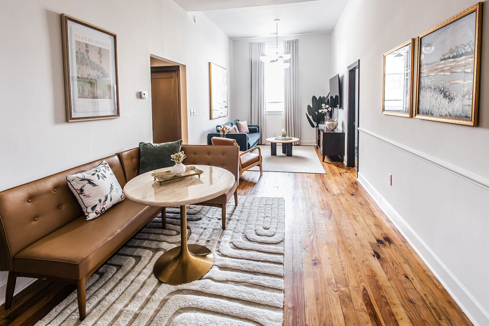 Un salon moderne avec un canapé en cuir marron, une table basse ronde et un plancher en bois. Des œuvres d’art encadrées ornent les murs, et une plante est posée sur la table d’appoint.