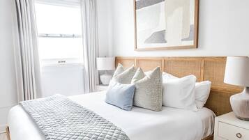Un dormitorio ordenado con cama, almohadas, una mesita de noche, lámpara, ventilador de techo y ventana con cortinas.