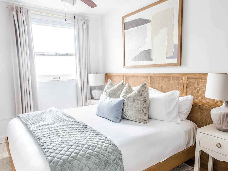 Un dormitorio ordenado con cama, almohadas, una mesita de noche, lámpara, ventilador de techo y ventana con cortinas.