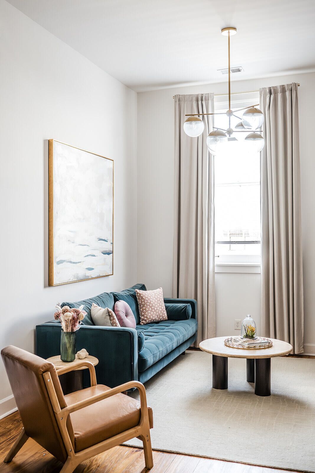 A modern living room with a blue sofa, a wooden chair, a round coffee table, and a large window with curtains.