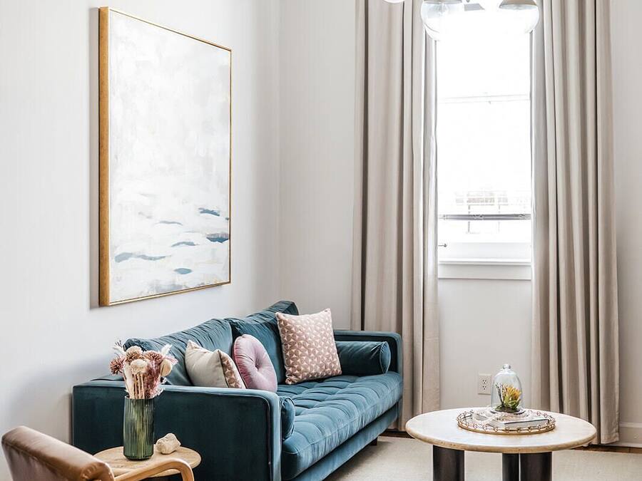 A modern living room with a blue sofa, a wooden chair, a round coffee table, and a large window with curtains.
