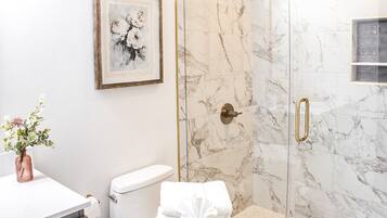 A bathroom with a toilet, a shower with glass doors, a marble wall, and a framed picture on the wall.