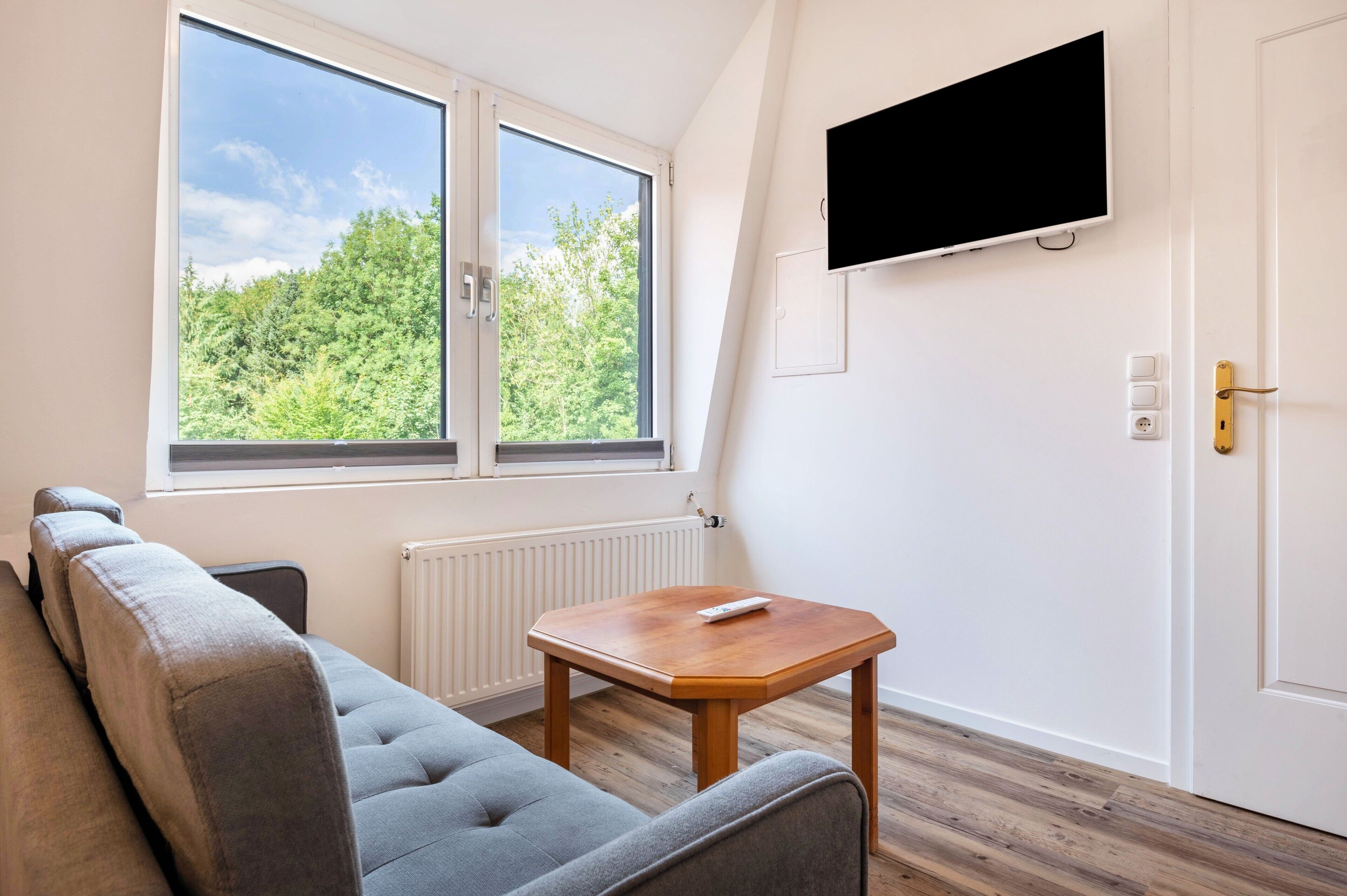 Ein Zimmer mit einer Couch, einem kleinen Tisch, einem an der Wand montierten Fernseher und einem Fenster mit Blick auf Bäume.