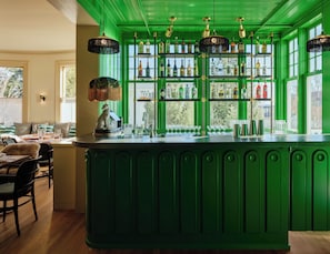 A green bar area with shelves of liquor bottles, a leopard statue, and a dining area with chairs and a table. - Belden House & Mews (Litchfield)