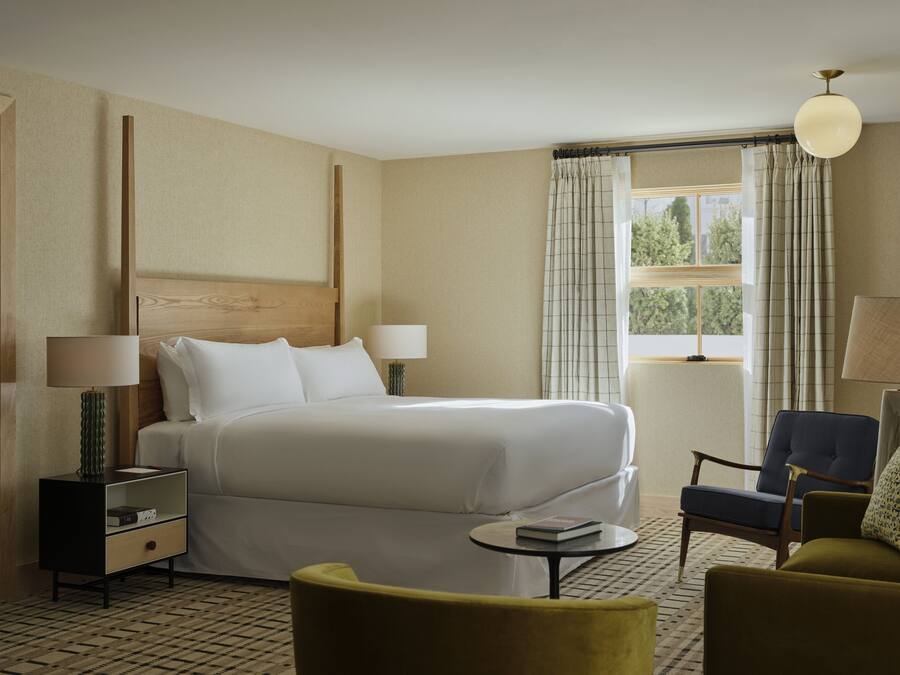 Quarto de hotel com cama, mesas de cabeceira, uma cadeira, uma mesinha e janela com cortinas.
