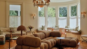 A spacious living room with a large sectional sofa, a round coffee table, and a chandelier. There are large windows with curtains, a fireplace, and a view of greenery outside.