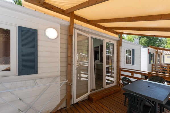 Uma área externa coberta com mesa e cadeiras, um deck de madeira e uma porta de vidro deslizante que dá para um espaço interno.