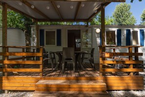 Een overdekt buitengebied met een houten terras, een tafel met stoelen en een klein gebouw met blauwe luiken.