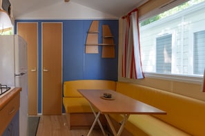 A compact kitchenette with a refrigerator, a sink, and a stove. There is a small table with a chair and a bench. The room has blue walls, wooden doors, and a window with curtains.