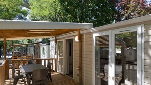 A covered outdoor deck with a table and chairs, a wooden pergola, and sliding glass doors leading to a covered patio area. - Mobile Home 'Confort B - 1-6 Pers' with Shared Pool, Private Terrace and Shared Garden (Lavilledieu)