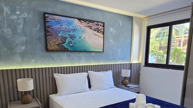 Ein Hotelzimmer mit einem großen Bett, einem Nachttisch, einer Lampe, einem Gemälde an der Wand und einem Fenster mit Blick auf Grünfläche.