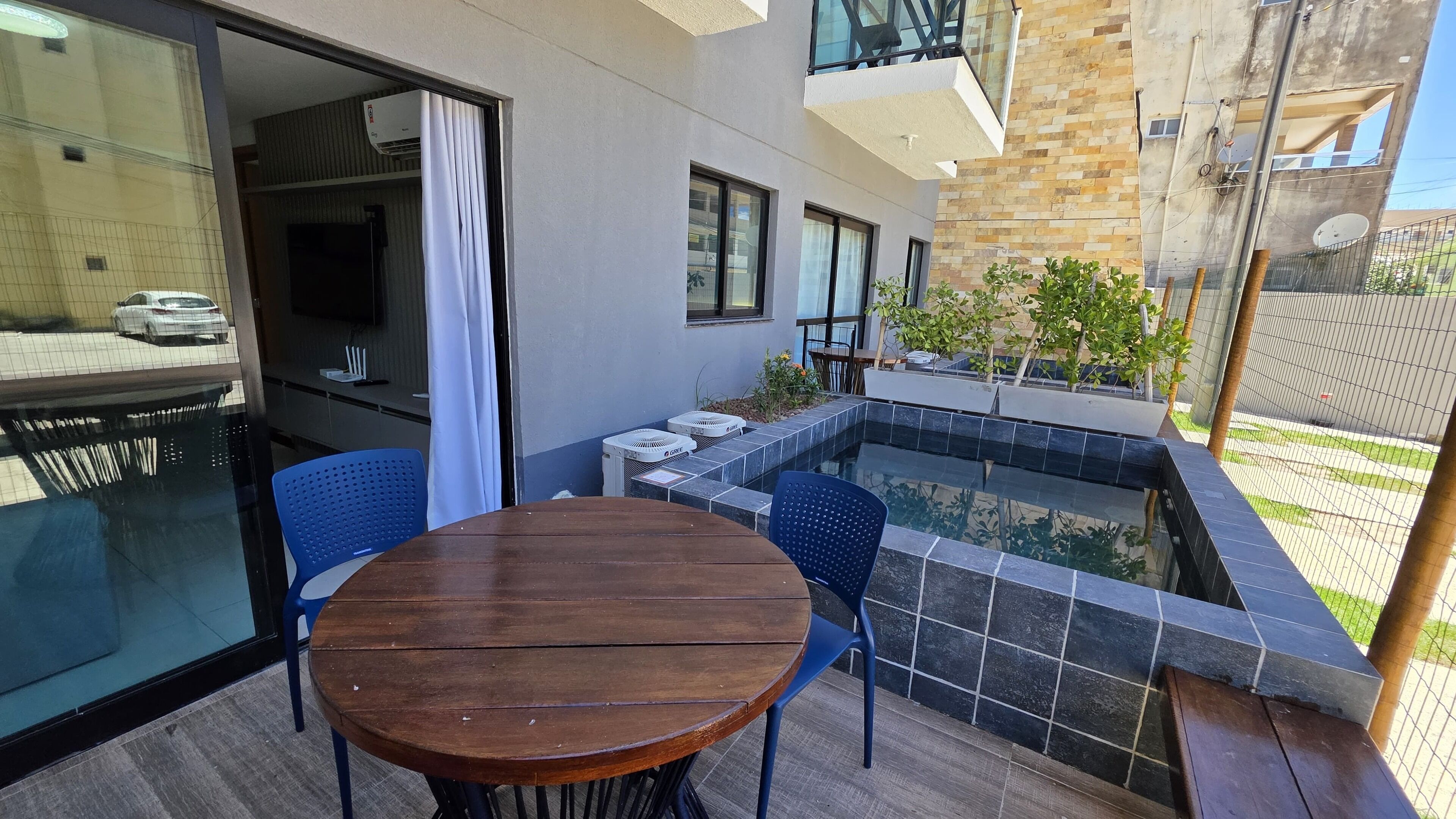 Uma área externa com mesa redonda de madeira e cadeiras azuis, uma piscina pequena com bordas azulejadas e vista para um edifício com grandes janelas e varanda.