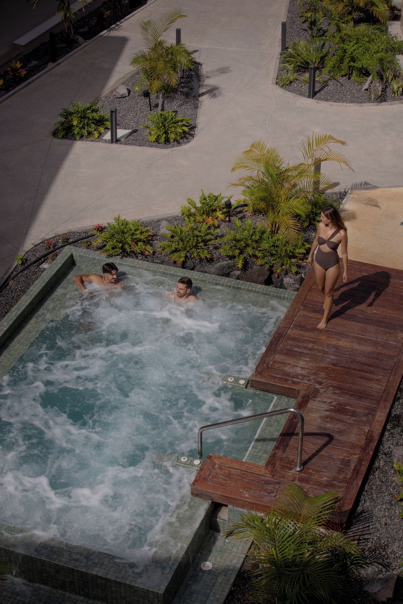 A hot tub area with a wooden deck, surrounded by landscaped gardens and a pool.