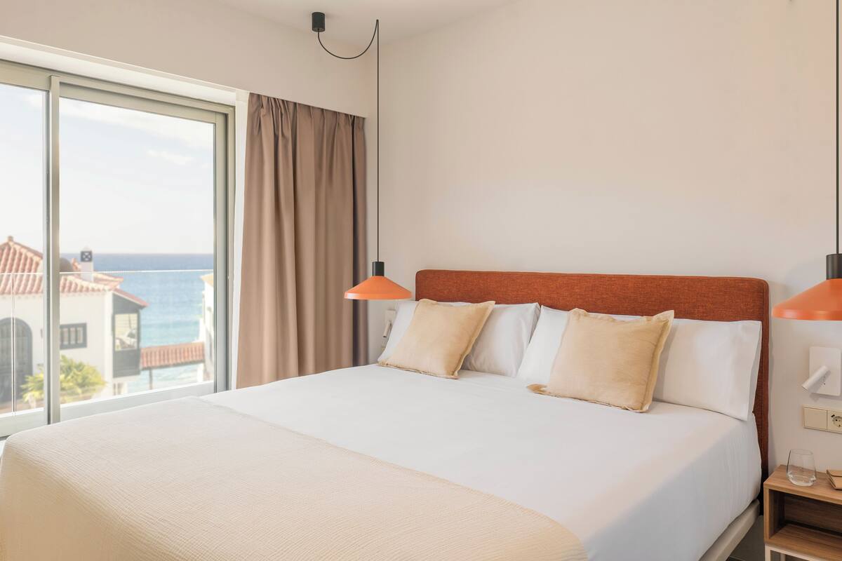 A hotel room with a large bed, two bedside lamps, a nightstand, and a view of the sea through a large window.