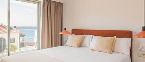 A hotel room with a large bed, two bedside lamps, a nightstand, and a view of the sea through a large window.