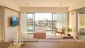 A hotel room with a large window, a flat-screen TV, a wooden table with a bottle and glass, a striped ottoman, and a chair. - KORA TIGOT (San Miguel de Abona)